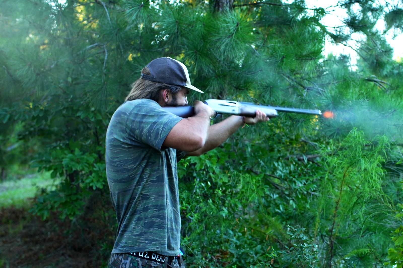 Turner firing a shotgun through the pines, muzzle flash visible